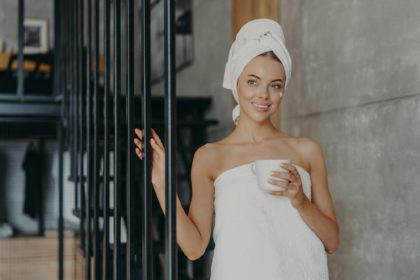 Healthy attractive woman with pensive expression smiles gently, applies face cream on complexion